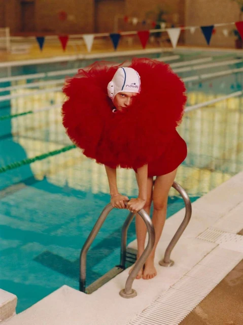 A woman in a red, flouncy dress and white swim cap poses by a blue indoor swimming pool, holding onto the metal pool ladder.