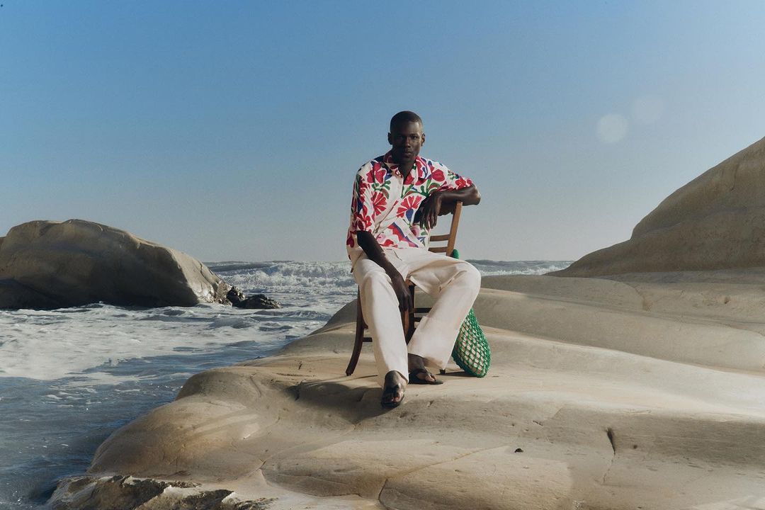 Man in floral shirt poses on rocky beach sitting on wooden chair with the ocean and sky visible in the background on a bright day.