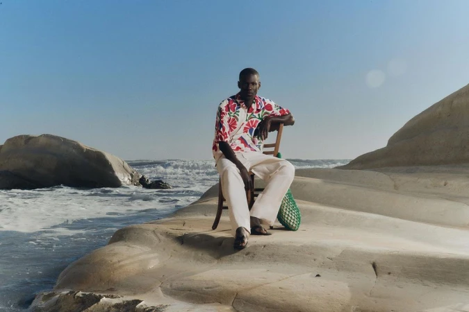 Man in patterned floral shirt and white trousers seated on a wooden chair on a pale rocky coastal formation near the ocean.