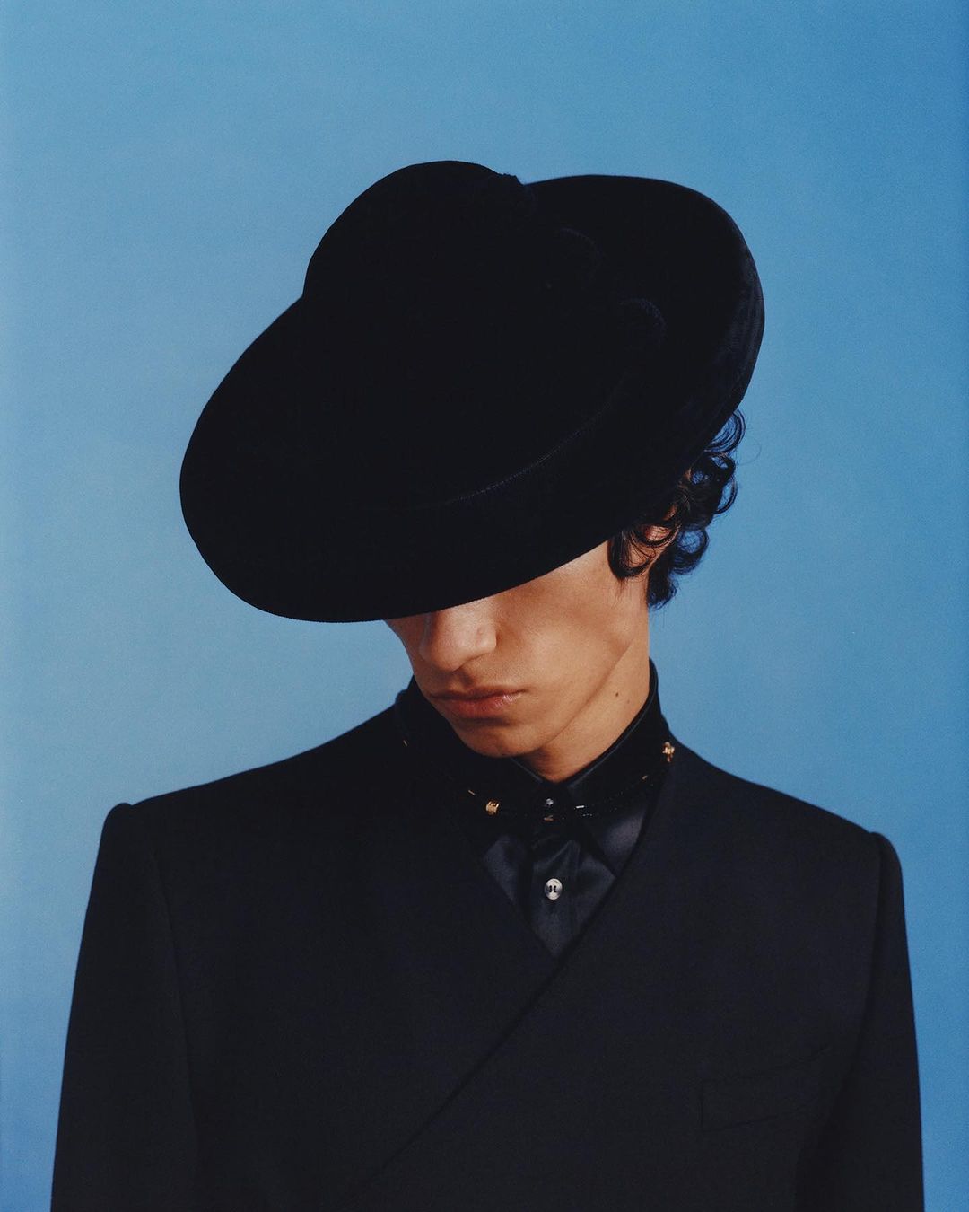 Close-up studio shot of androgynous adult in a black suit and wide-brimmed hat, set against a simple blue backdrop in portrait orientation.