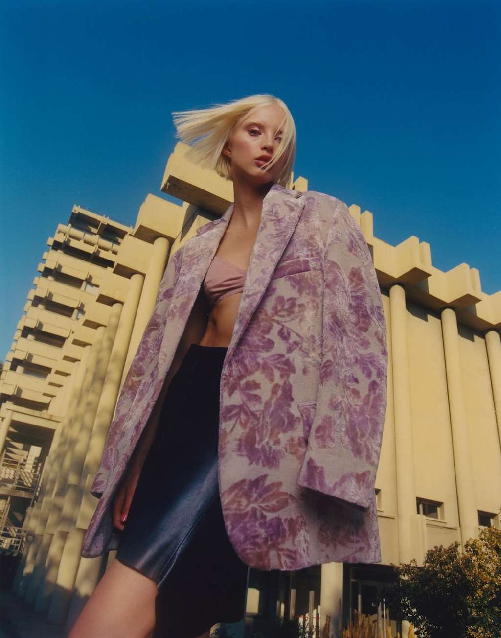 A woman in a floral blazer stands against a blue sky and a geometric building, creating a fashion-editorial image with architectural backdrop.