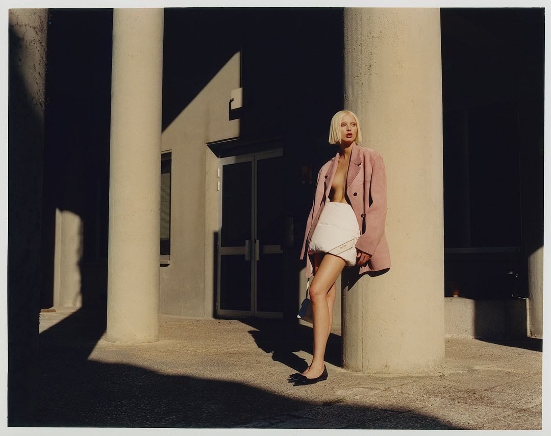 Young woman with platinum bob poses in a pink blazer and skirt in an outdoor setting, leaning against a concrete pillar, fashion editorial.
