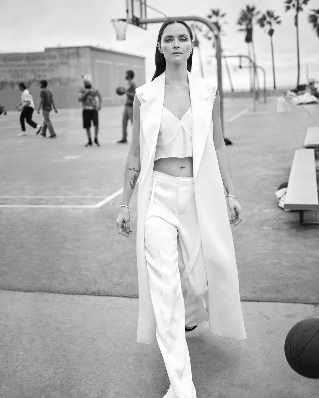 A woman in a white outfit stands on an outdoor basketball court, wearing a sleeveless long jacket with children playing in the background.
