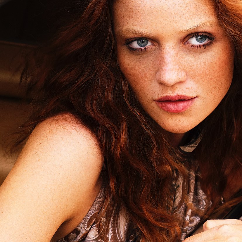 Close-up portrait of a red-haired, light-skinned adult female model with freckles and blue eyes looking directly at the camera indoors.