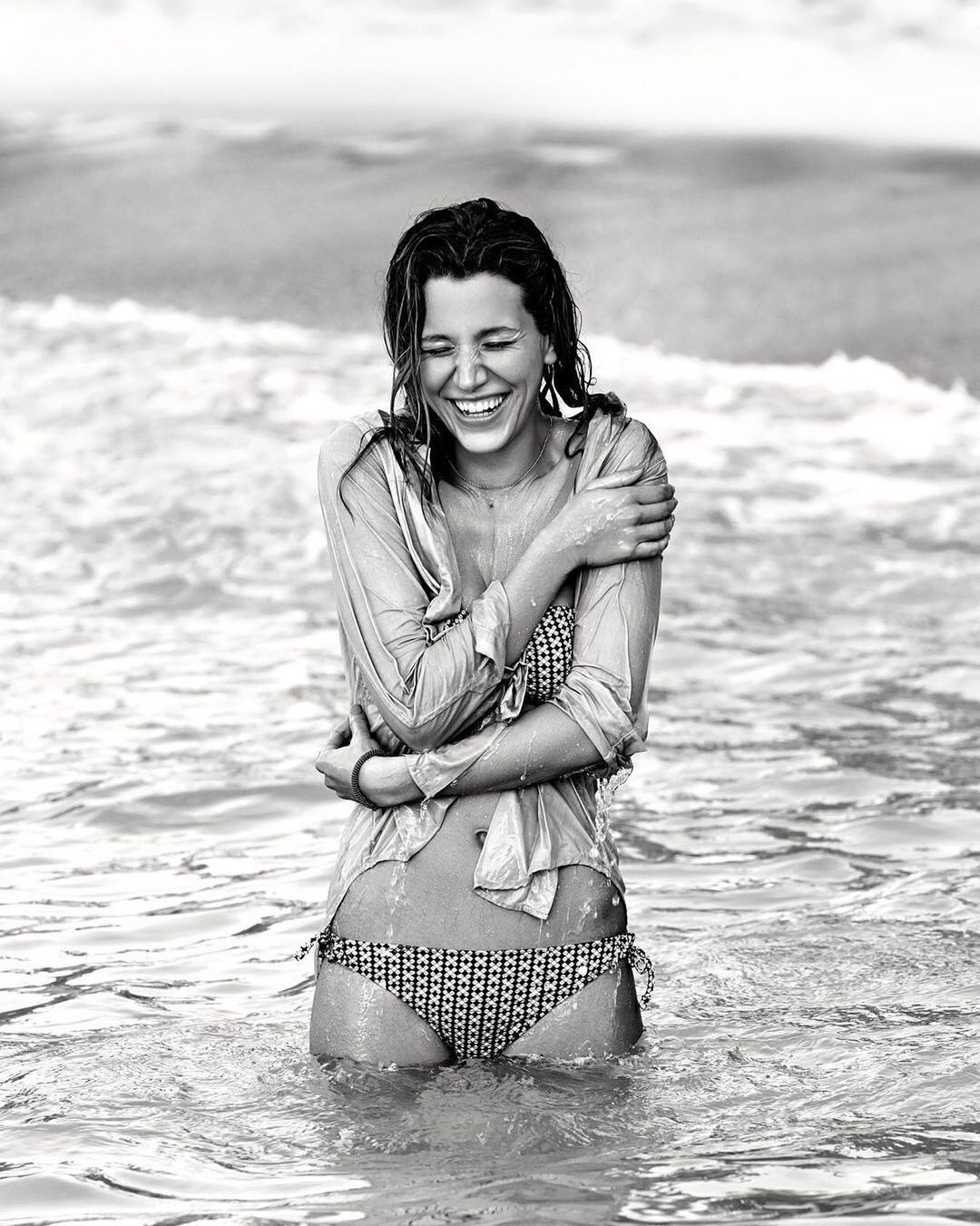 Monochrome portrait of a smiling young woman standing in the ocean surf wearing a patterned bikini and wet shirt with her arms crossed.