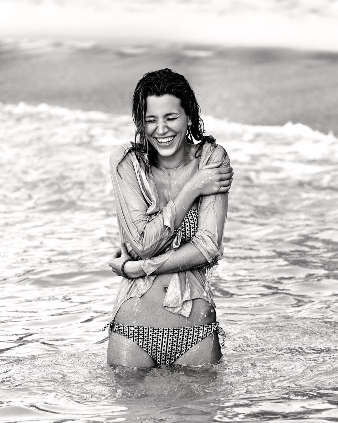 Monochrome shot of a smiling woman in a bikini and shirt standing in ocean waves, wet hair, and arms crossed, exuding happiness.