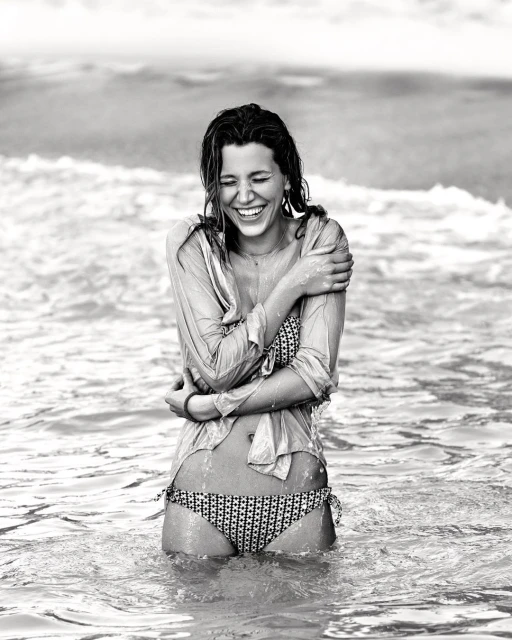 Monochrome shot of a smiling woman in a bikini and shirt standing in ocean waves, wet hair, and arms crossed, exuding happiness.