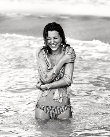 Monochrome shot of a smiling woman in a bikini and shirt standing in ocean waves, wet hair, and arms crossed, exuding happiness.