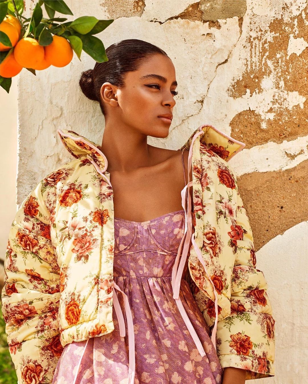 Stylish woman in a floral jacket and dress poses outdoors with oranges and an aged wall, bathed in soft natural light.