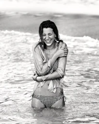 A smiling woman wearing a patterned bikini and a wet button-down shirt standing in ocean surf