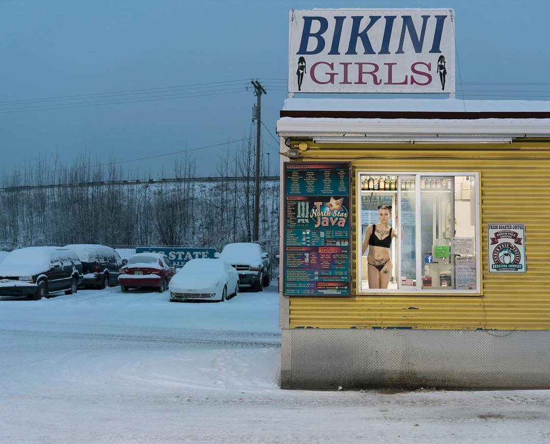 Woman in bikini inside a yellow coffee stand called Bikini Girls during winter with snow-covered cars, trees, and ground.