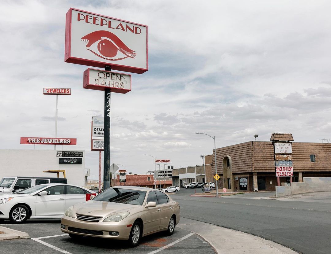 Roadside view of a Peepland adult store sign on a pole with a parked car in front of the building, several businesses are nearby.