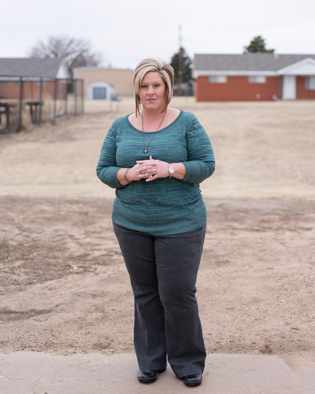 Portrait of a middle-aged Caucasian woman standing in an outdoor field with buildings in the background under an overcast sky.