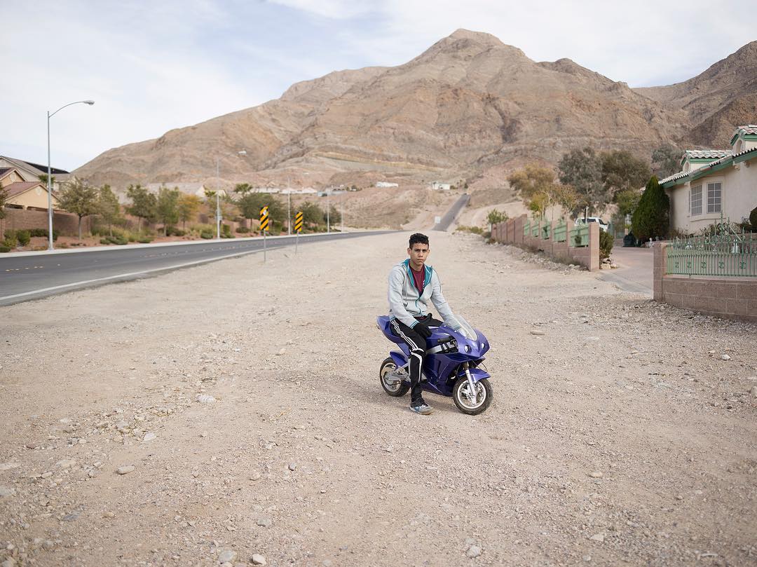 A young man sits on a blue toy motorcycle on a dirt patch, with a road, houses, and a tan mountain in the background on a sunny day.