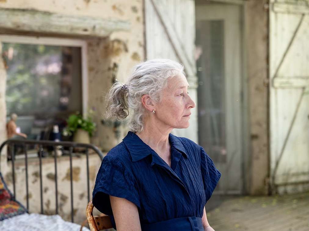 Mature woman with silver hair sits outside a stone building, wearing a navy blue dress in a medium shot with natural daylight.