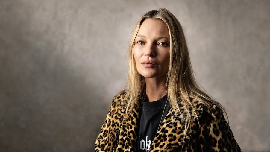Portrait of Kate Moss wearing a leopard-print coat and black T-shirt, set against a textured grey background in soft lighting.