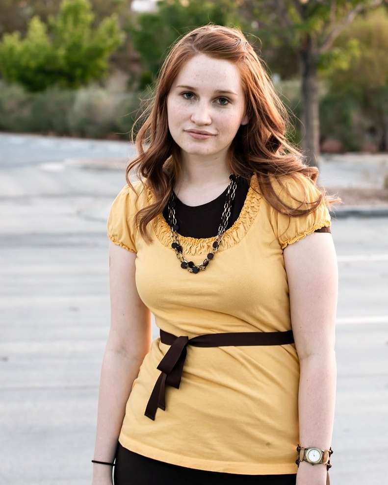 Young woman poses outdoors wearing a yellow top with a brown belt and black skirt in an eye-level medium shot with blurred background.