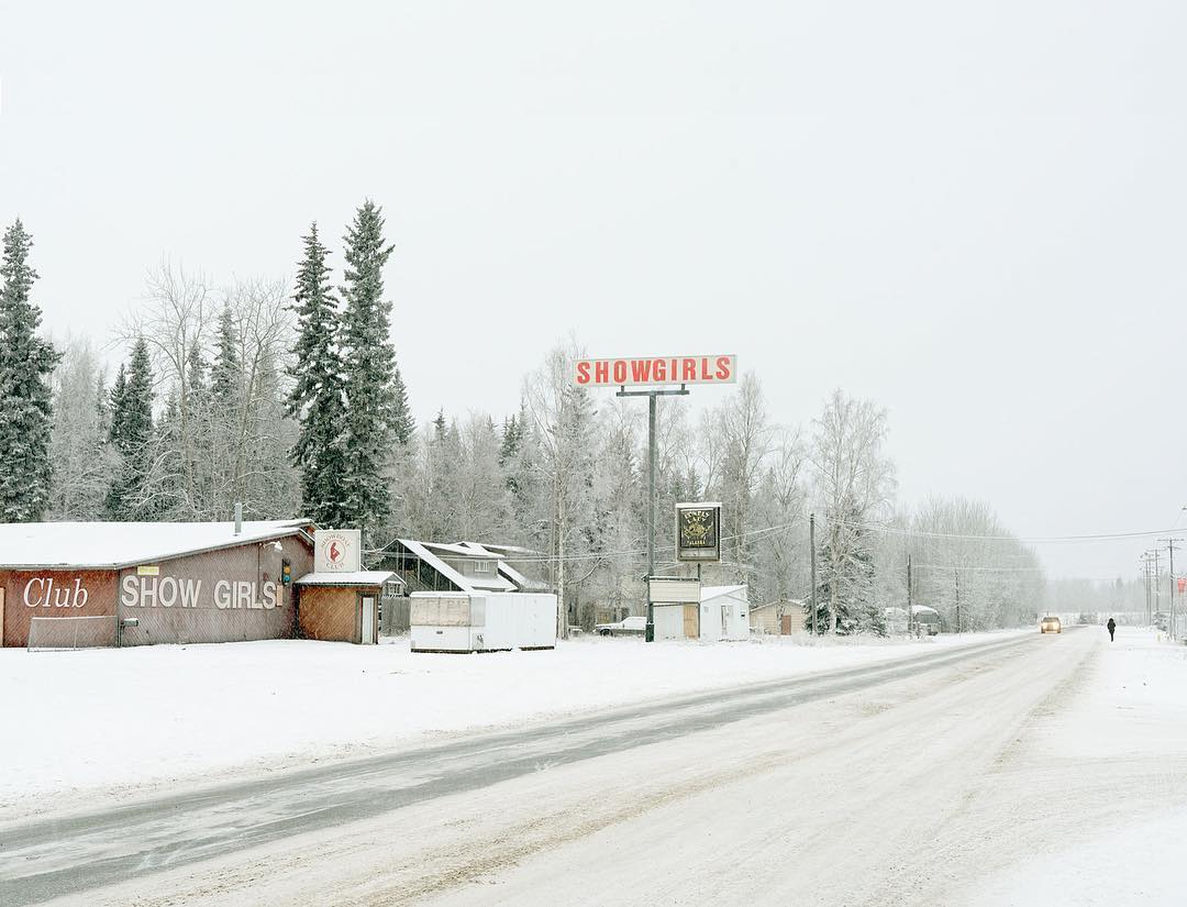 Snowy rural scene featuring a 'Showgirls' sign and club, with sparse trees and a distant figure walking along the snow-covered road.