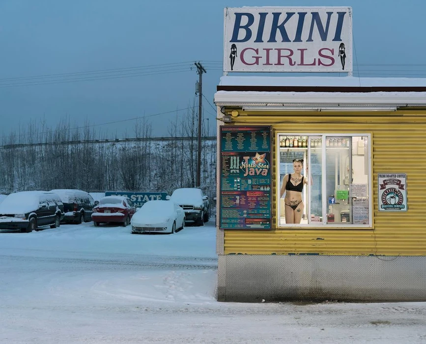 Woman in black lingerie standing in a lit kiosk window with a Bikini Girls sign over a snowy parking lot