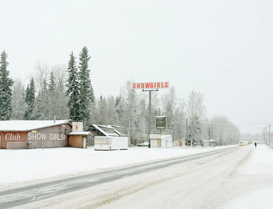 Snow-covered roadside scene featuring a building with Show Girls signage and tall pine trees under an overcast sky.