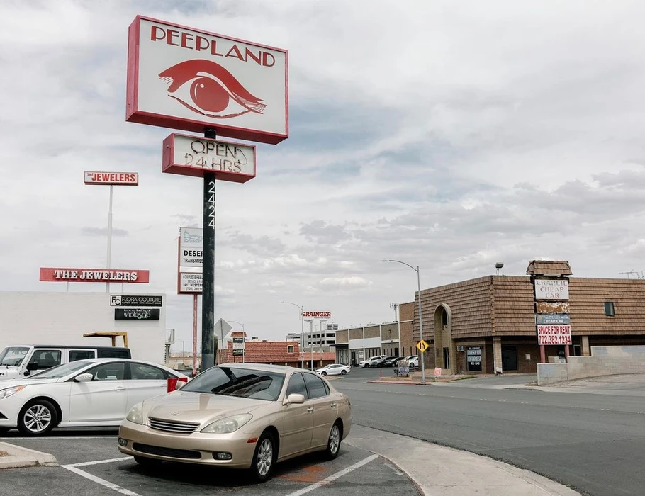 Parked gold sedan and white car under a large Peepland sign on an overcast day in Las Vegas.