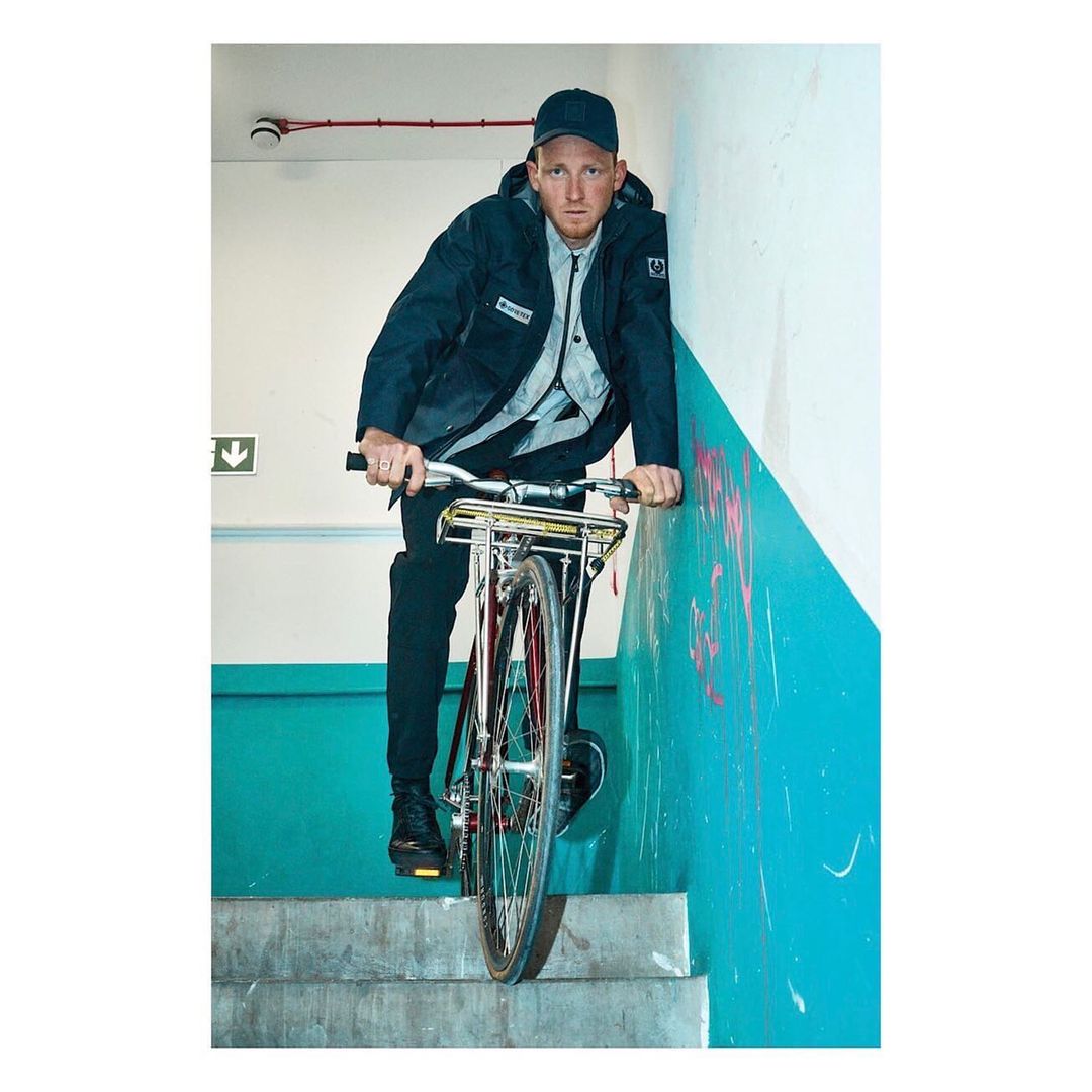 A man in a dark jacket rides a red bicycle down stairs in an indoor location with teal-and-white walls, looking directly at the camera.