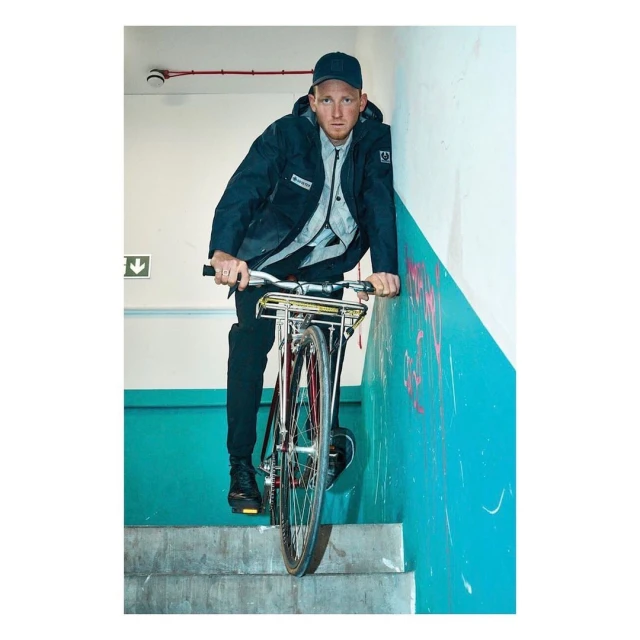 A man in a dark jacket rides a red bicycle down stairs in an indoor location with teal-and-white walls, looking directly at the camera.