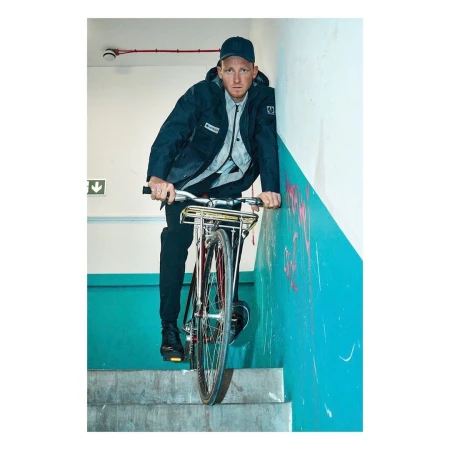 A man in a dark jacket rides a red bicycle down stairs in an indoor location with teal-and-white walls, looking directly at the camera.