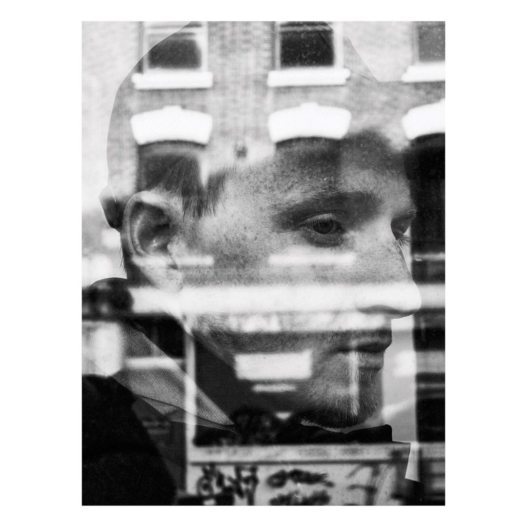 Monochrome portrait of a young man with a building reflection creating a double exposure effect, rendered in black and white with visible texture.
