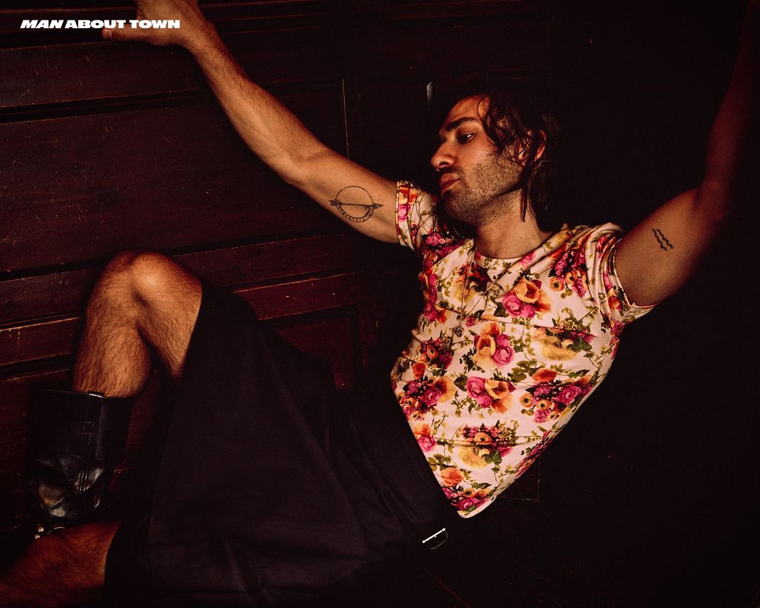 Man with floral t-shirt and tattoos posed against a dark wood wall, photographed by David Hughes in London.