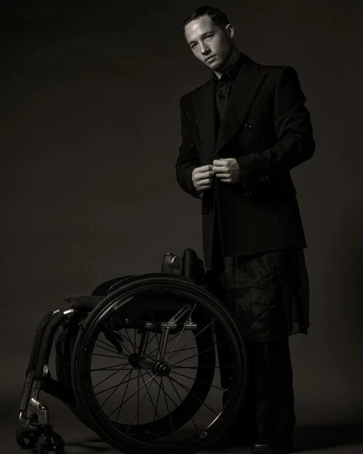 Stylish man in a dark suit standing next to a wheelchair in a studio setting. Fashion-forward and artistic portrait, suitable for editorial or personal profile.