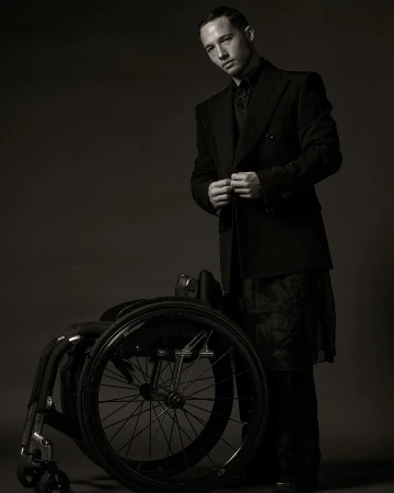 Stylish man in a dark suit standing next to a wheelchair in a studio setting. Fashion-forward and artistic portrait, suitable for editorial or personal profile.