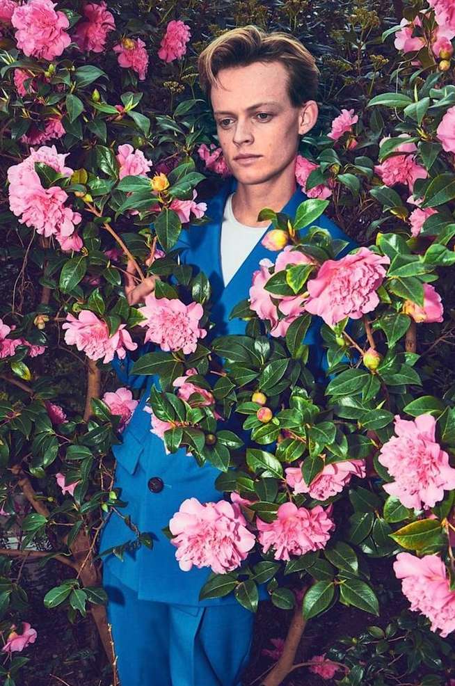 Portrait of a blond male model wearing a blue suit, surrounded by pink camellia flowers and green leaves in an outdoor setting.
