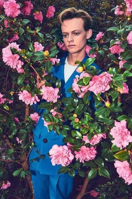 Portrait of a blond male model wearing a blue suit, surrounded by pink camellia flowers and green leaves in an outdoor setting.