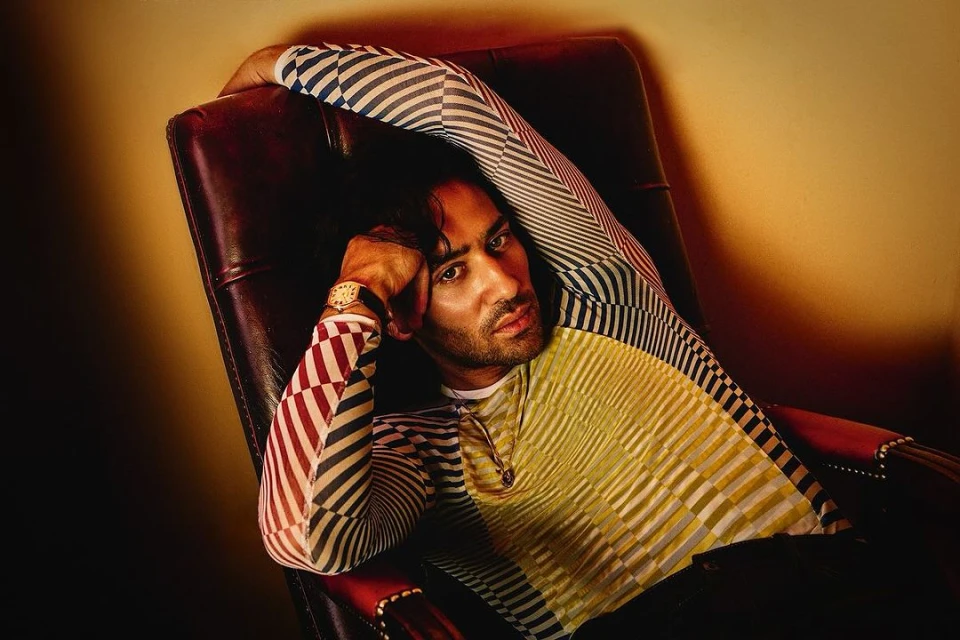 Man in patterned shirt reclines in a leather armchair. Indoor studio portrait with warm lighting and a confident expression.