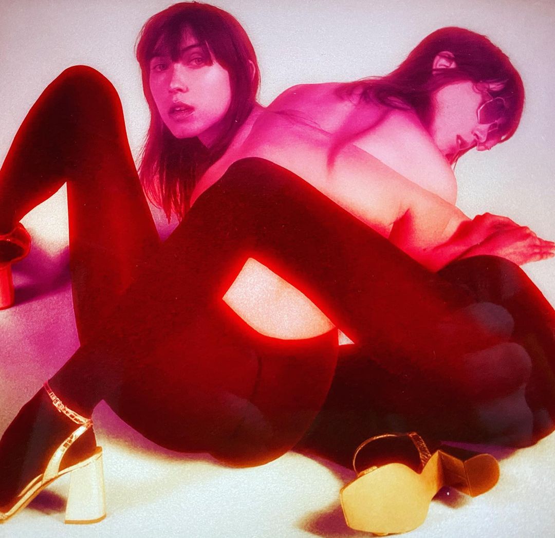 Two women pose in a photo studio. One woman sits with her legs extended. The other lays behind her with sunglasses. Red and pink tones.