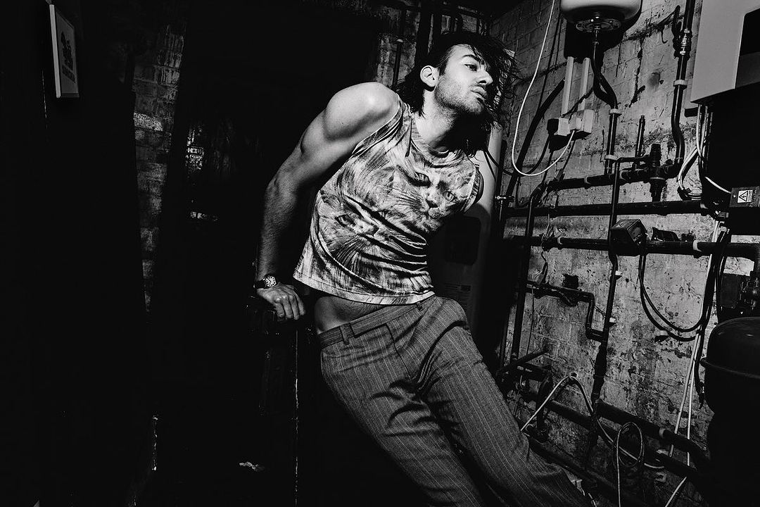 Black-and-white portrait of a muscular adult male wearing a printed tank top and pinstripe pants, posing in front of old plumbing.