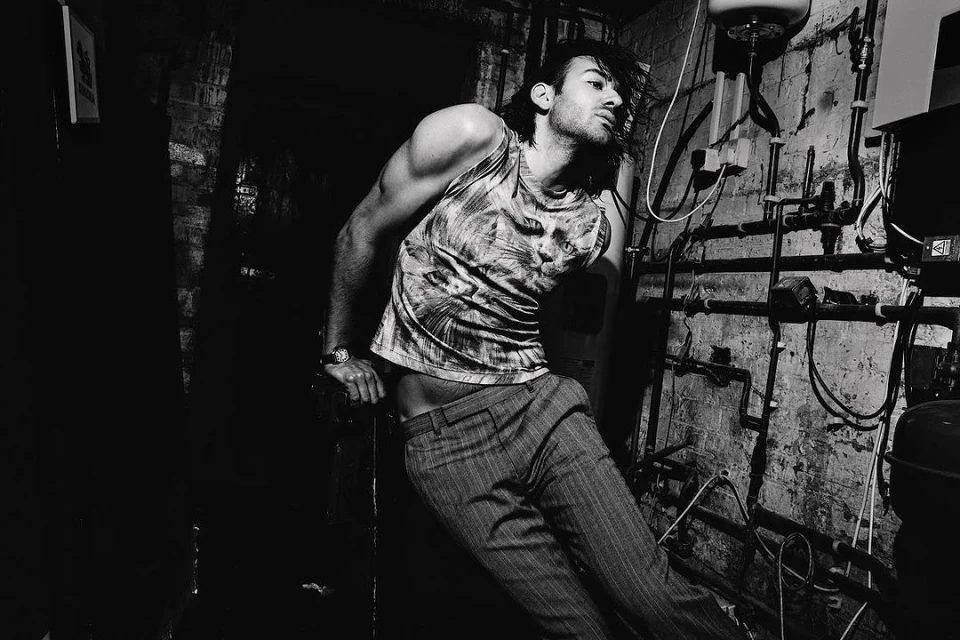 Black-and-white portrait of a muscular adult male wearing a printed tank top and pinstripe pants, posing in front of old plumbing.