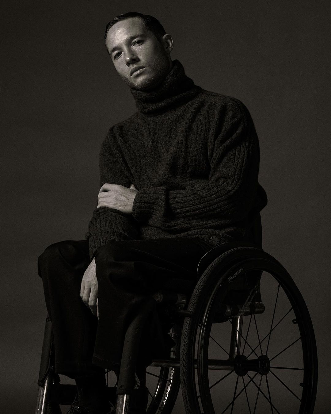 Portrait of an adult male with short hair wearing a dark turtleneck sweater, sitting in a wheelchair against a dark grey background.