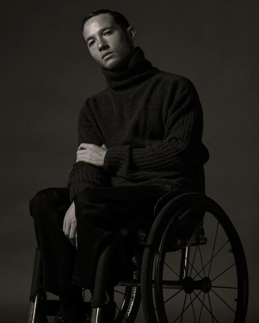Portrait of an adult male with short hair wearing a dark turtleneck sweater, sitting in a wheelchair against a dark grey background.