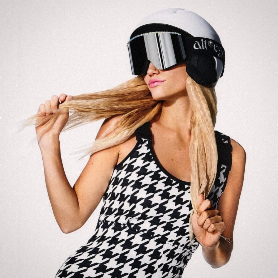 A fair-skinned woman with long blonde hair, wearing a white helmet and goggles, poses against a white background in a houndstooth dress.