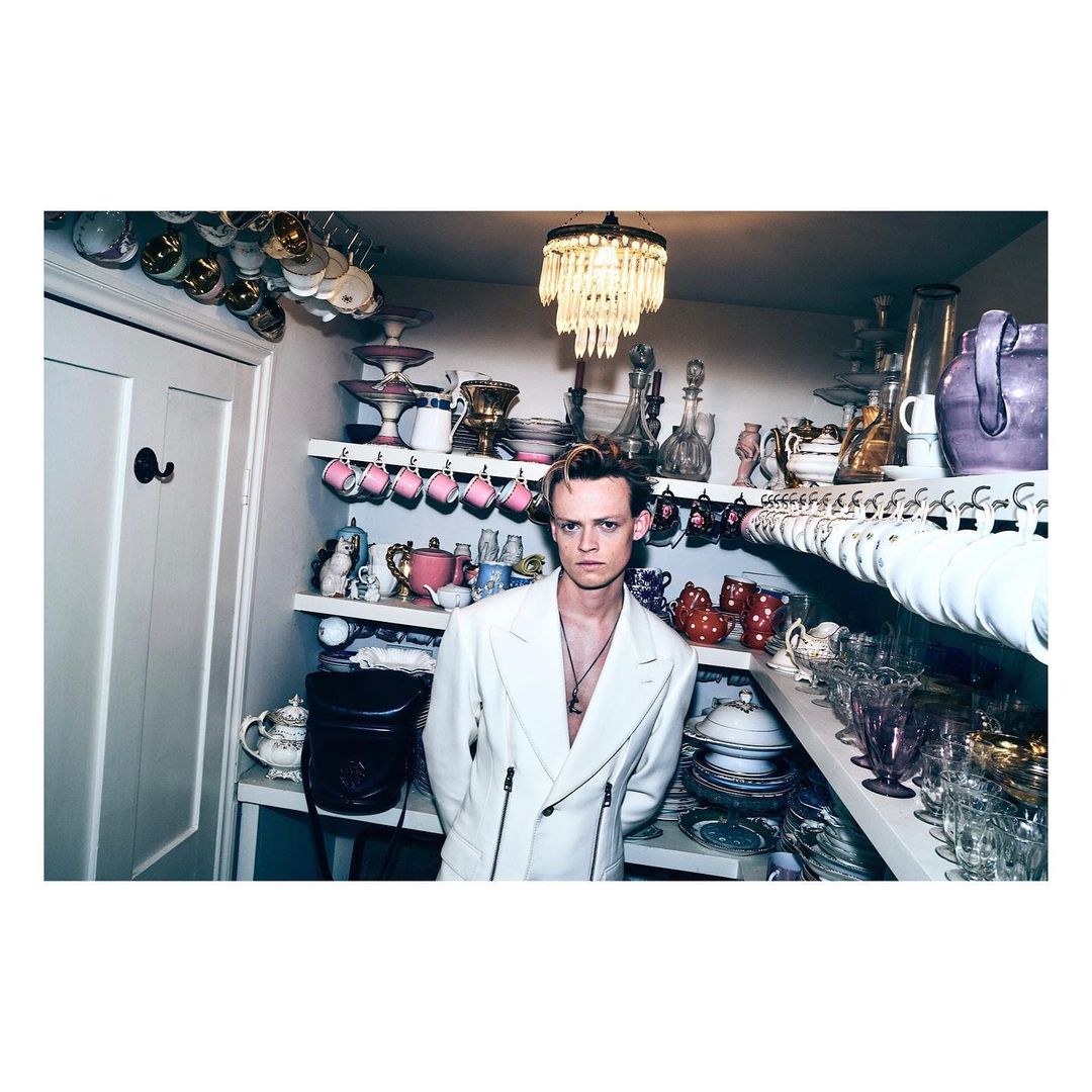 Portrait of a slender adult male wearing a white blazer in an indoor kitchen setting with shelves of antique dishware.