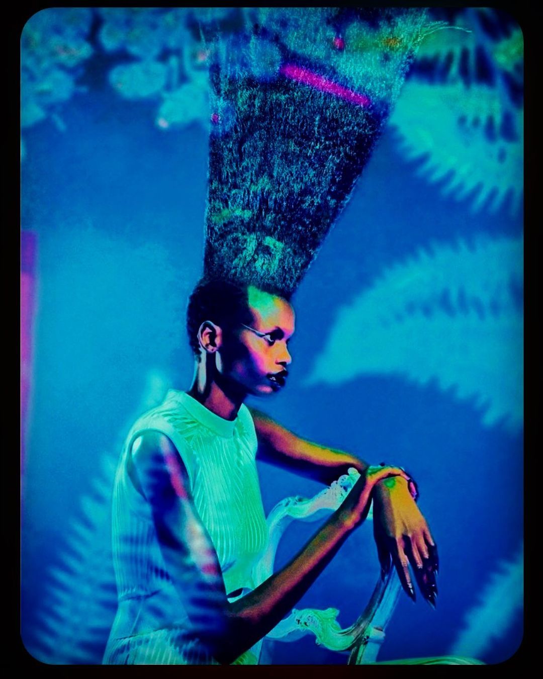 Striking fashion shot of a black female model with a sky-high beehive hairdo, wearing sleeveless top, and sitting on a decorative chair.