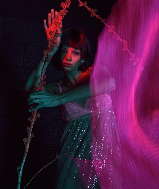 Woman in sequin dress poses in a dimly lit indoor setting with dark walls, illuminated by pink and green lighting.