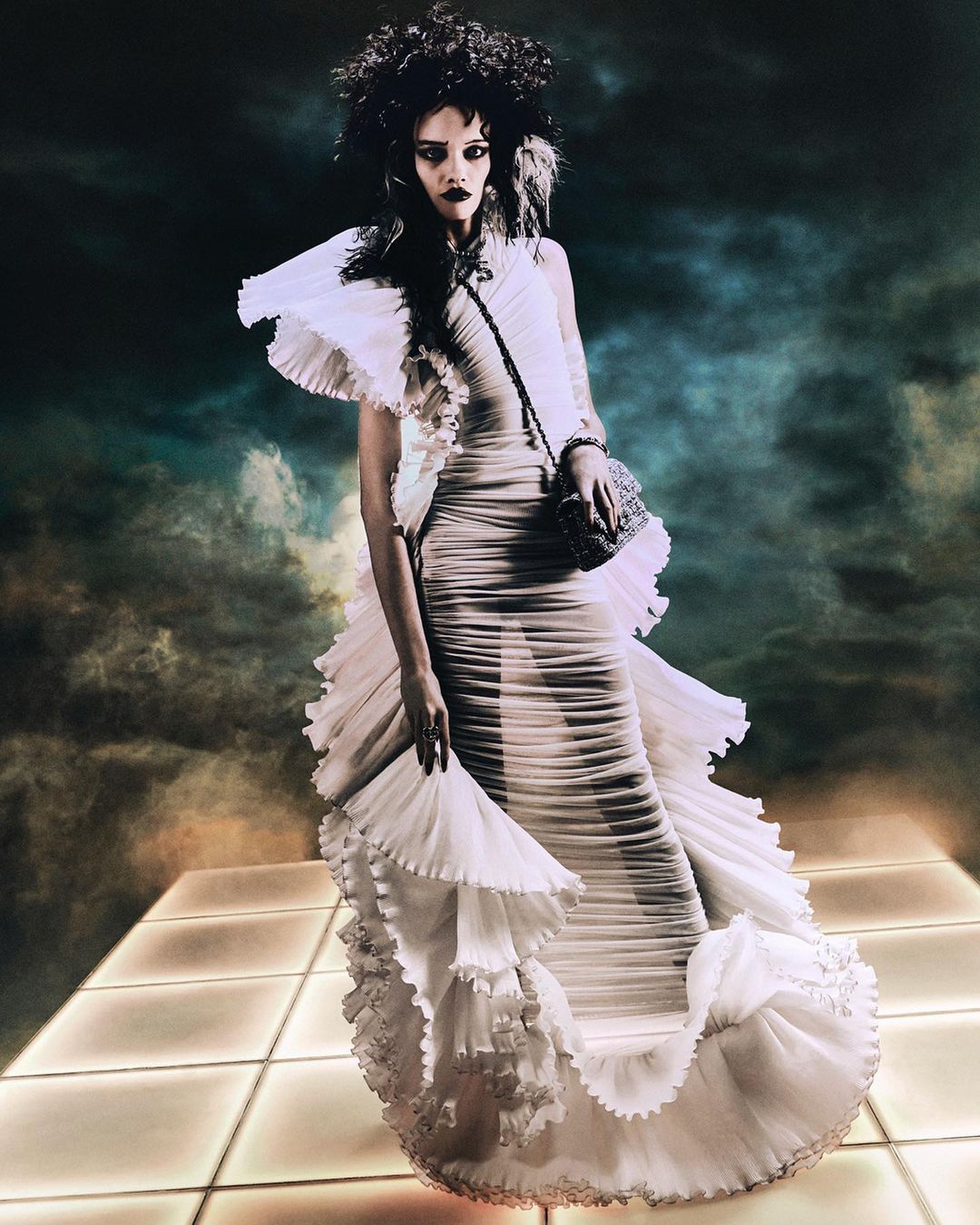 Model wearing a pleated white gown with dramatic ruffles standing on a lit grid platform under a dramatic stormy sky.