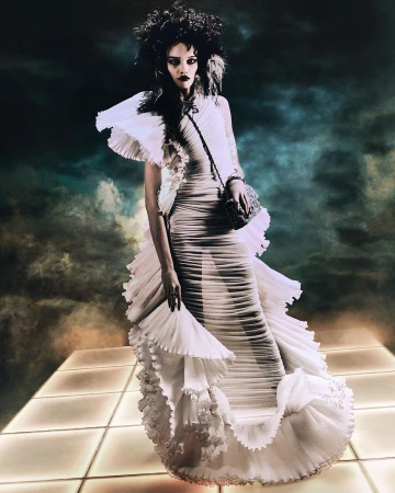 Model wearing a pleated white gown with dramatic ruffles standing on a lit grid platform under a dramatic stormy sky.