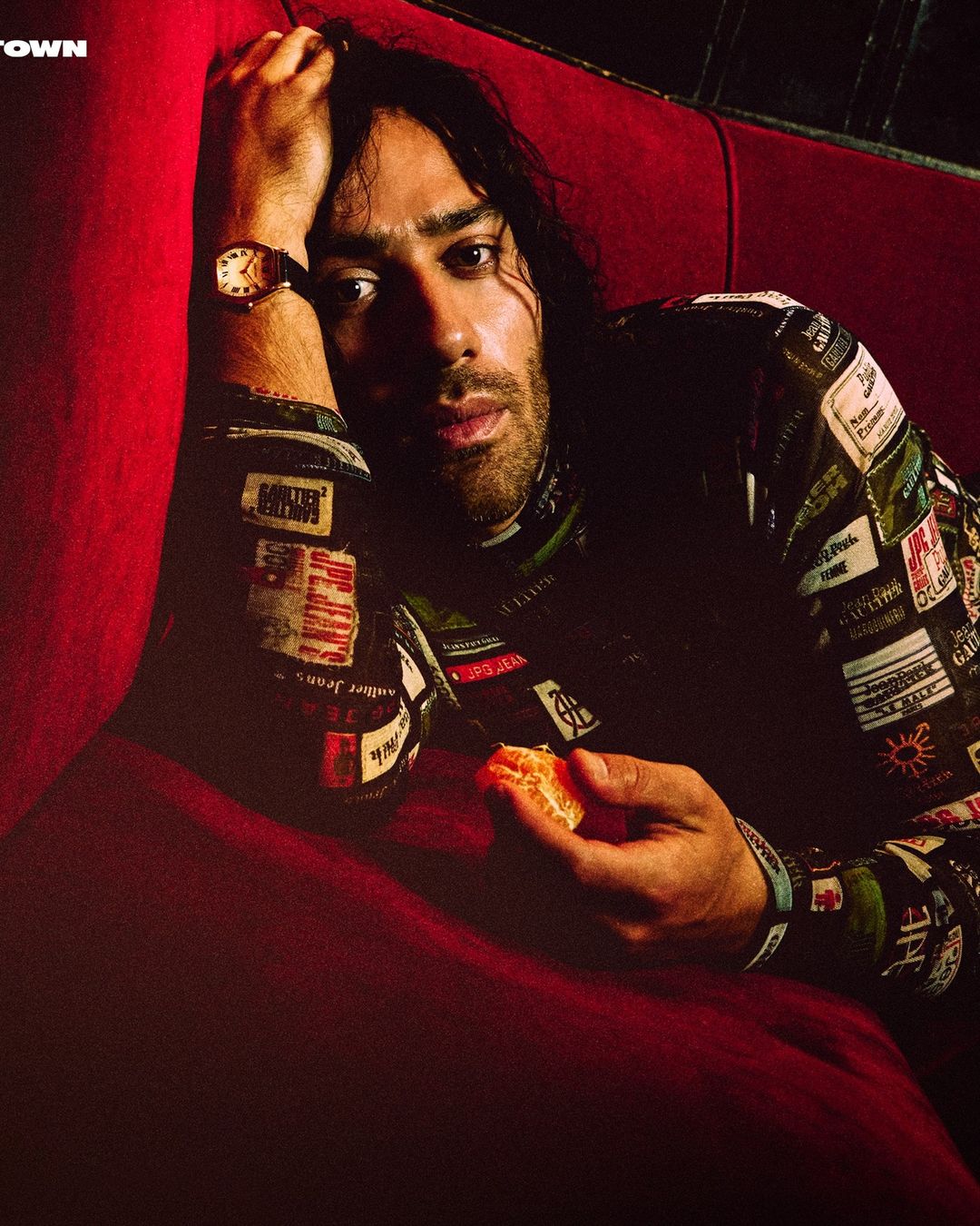 Portrait of a man with long hair wearing a patch jacket, reclining on a red sofa with a relaxed expression and holding citrus.