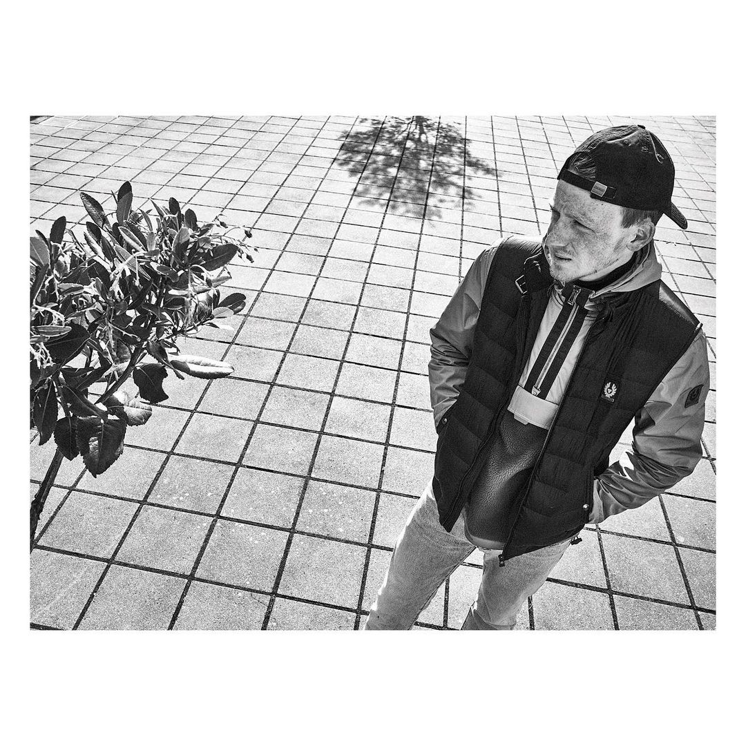 Monochrome portrait of a young man wearing a cap and vest standing on a tiled surface, with a plant and tree shadow visible.