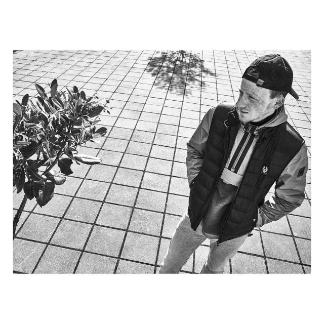 Monochrome portrait of a young man wearing a cap and vest standing on a tiled surface, with a plant and tree shadow visible.