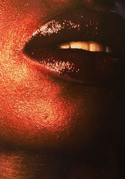 Extreme close-up captures a person's face, with dark, glossy lips as the focal point, set against warm-toned, textured skin.